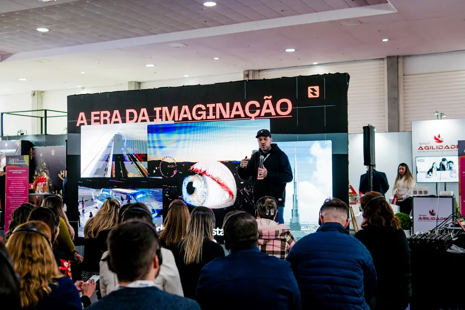 Gui Zanoni de boné e moletom, com o microfone na mão, numa palestra sobre IA num palco de feira. No telão, o título A Era da Imaginação. Na frente, a plateia ouve com fones de ouvido.