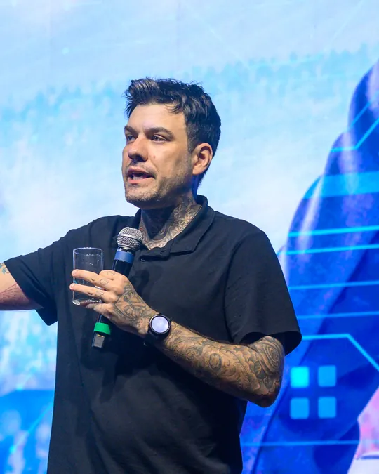 Gui Zanoni no palco, de camisa polo preta, com um copo de água e o microfone na mesma mão, respondendo à plateia.