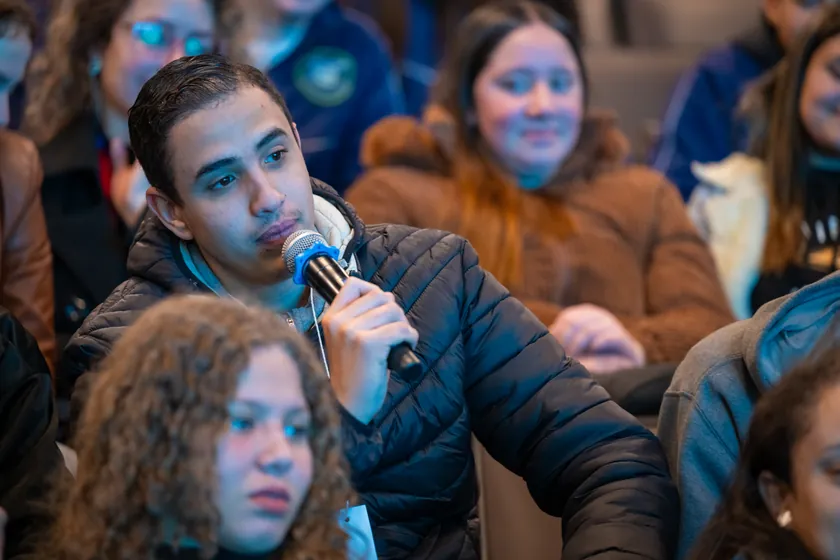 Um rapaz de jaqueta acolchoada faz uma pergunta ao microfone, sentado no meio da plateia. Em volta, outras pessoas jovens ouvem.