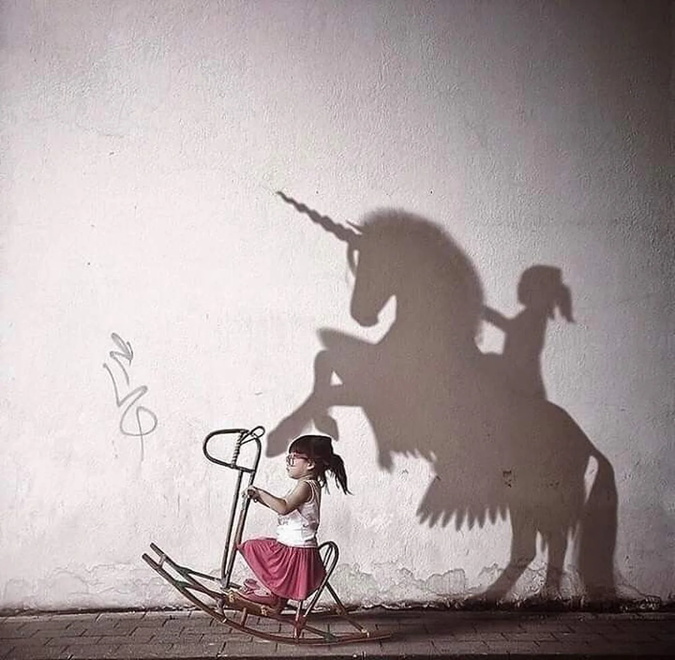 Uma menina num cavalinho de balanço, diante de uma parede branca. A sombra dela na parede é a de alguém montando um unicórnio.