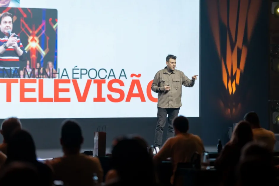 Gui Zanoni no palco, de camisa, apontando para a plateia. No telão atrás dele, a frase: na minha época, a televisão.