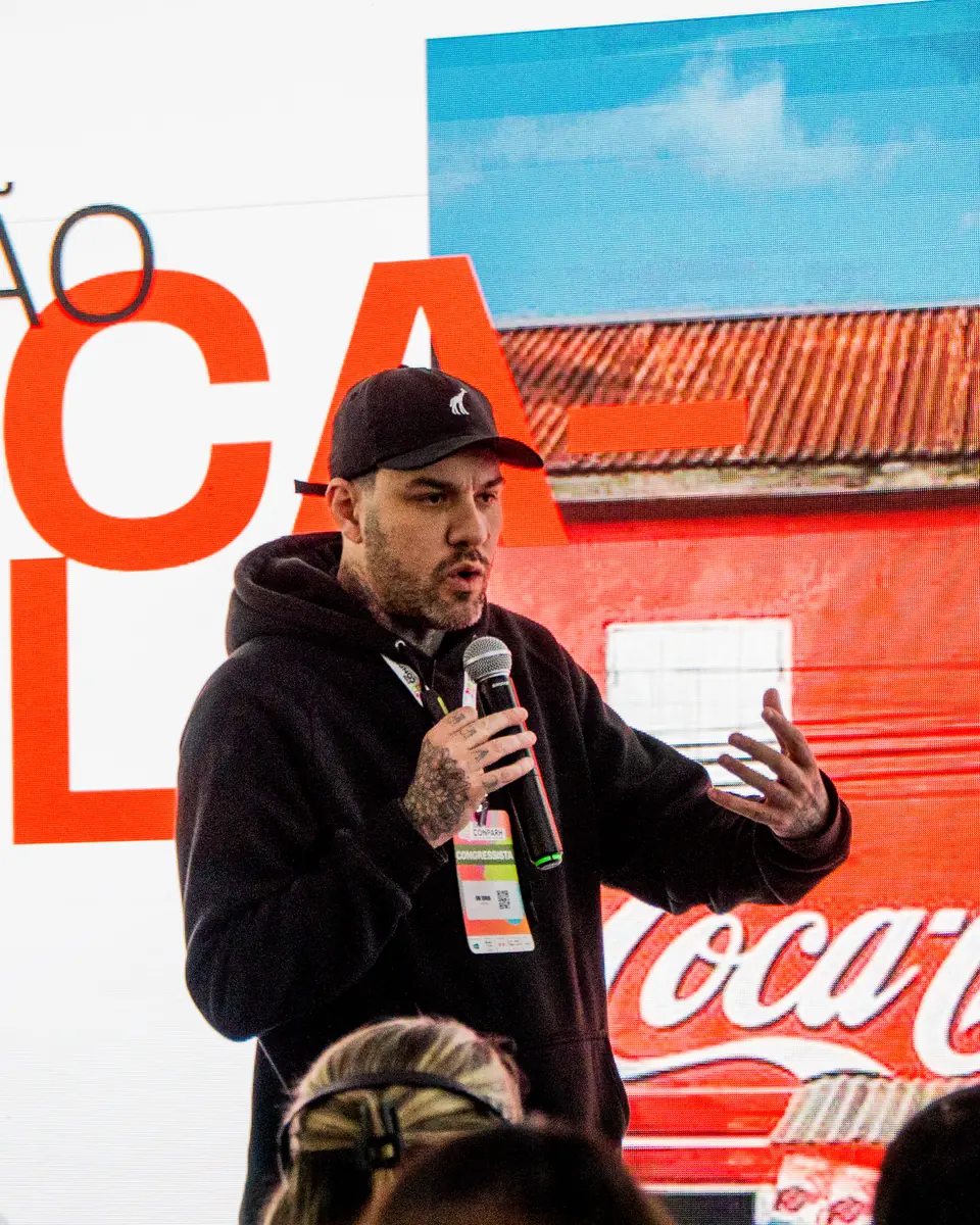 Gui Zanoni no palco do CONPARH, de boné e moletom pretos, falando ao microfone diante de um telão com o título Geração Coca-Cola e a fachada vermelha de um bar com a marca.