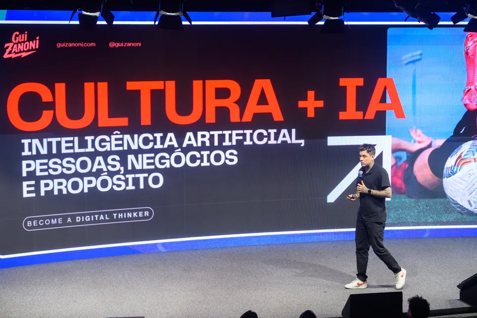 Gui Zanoni de camisa preta caminhando num palco grande. No telão atrás dele, em letras laranja: Cultura + IA. Inteligência artificial, pessoas, negócios e propósito.