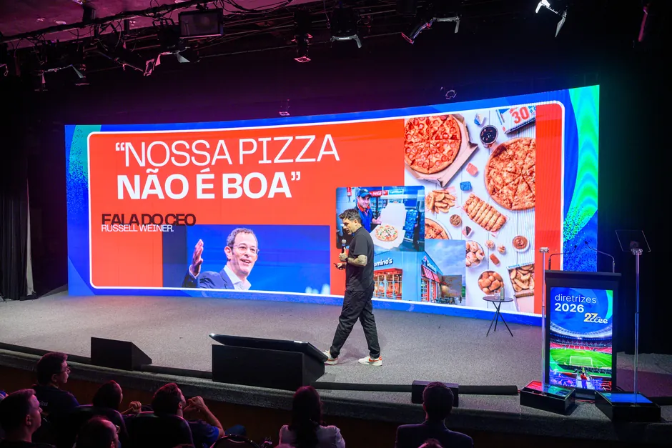 Gui Zanoni num palco de auditório diante de um telão curvo com a frase entre aspas: nossa pizza não é boa, fala do CEO Russell Weiner, com fotos de pizzas e de uma loja Domino's. Plateia de costas em primeiro plano.