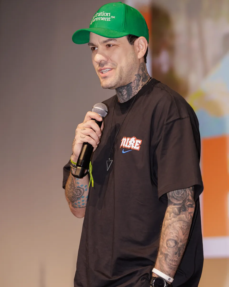 O palestrante futurista Gui Zanoni no palco, de boné verde e camiseta preta, falando ao microfone.