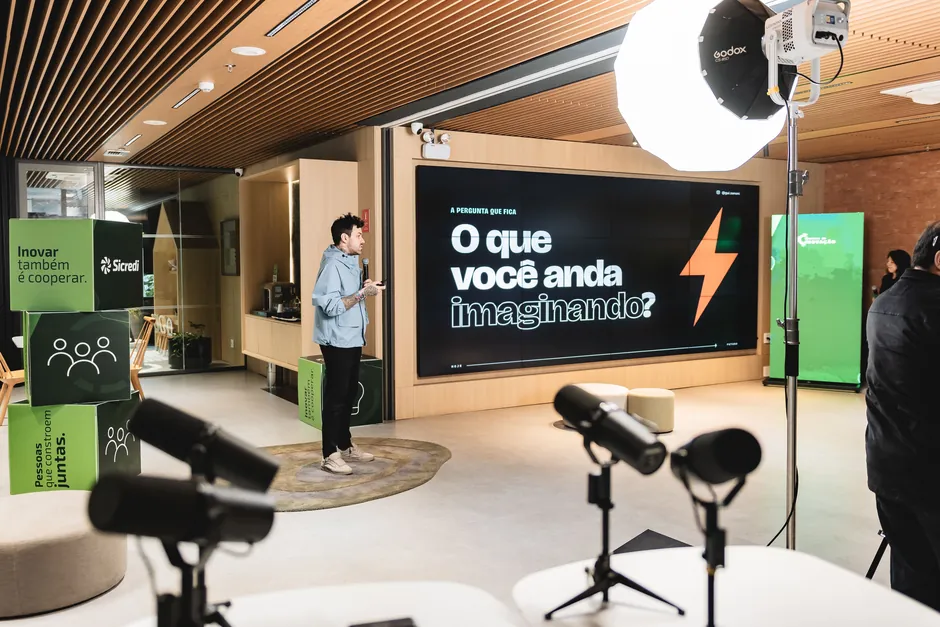 Gui Zanoni de jaqueta azul clara, com microfone, num espaço de inovação com cubos verdes do Sicredi. No telão, a pergunta: O que você anda imaginando?
