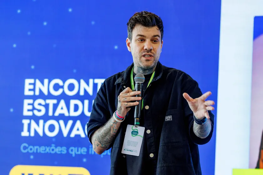 Gui Zanoni falando ao microfone, de camisa escura e crachá, diante de um painel azul com o título Encontro Estadual de Inovação.