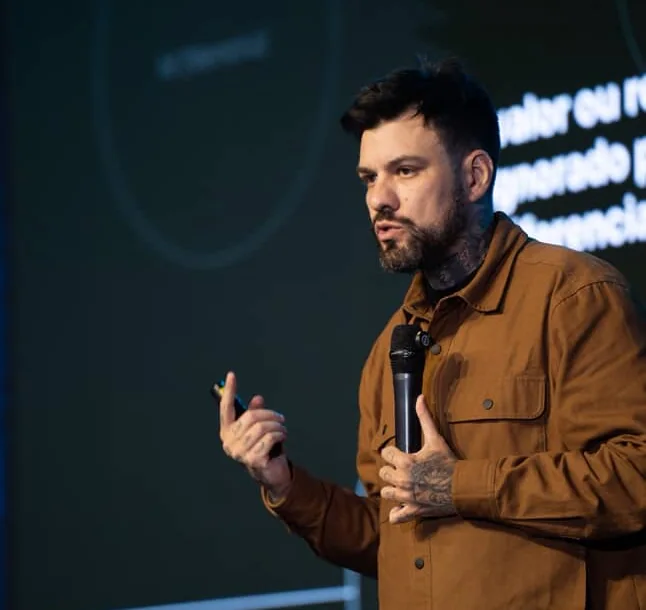 Gui Zanoni de camisa marrom, com microfone e passador de slides, num palco escuro diante do telão.