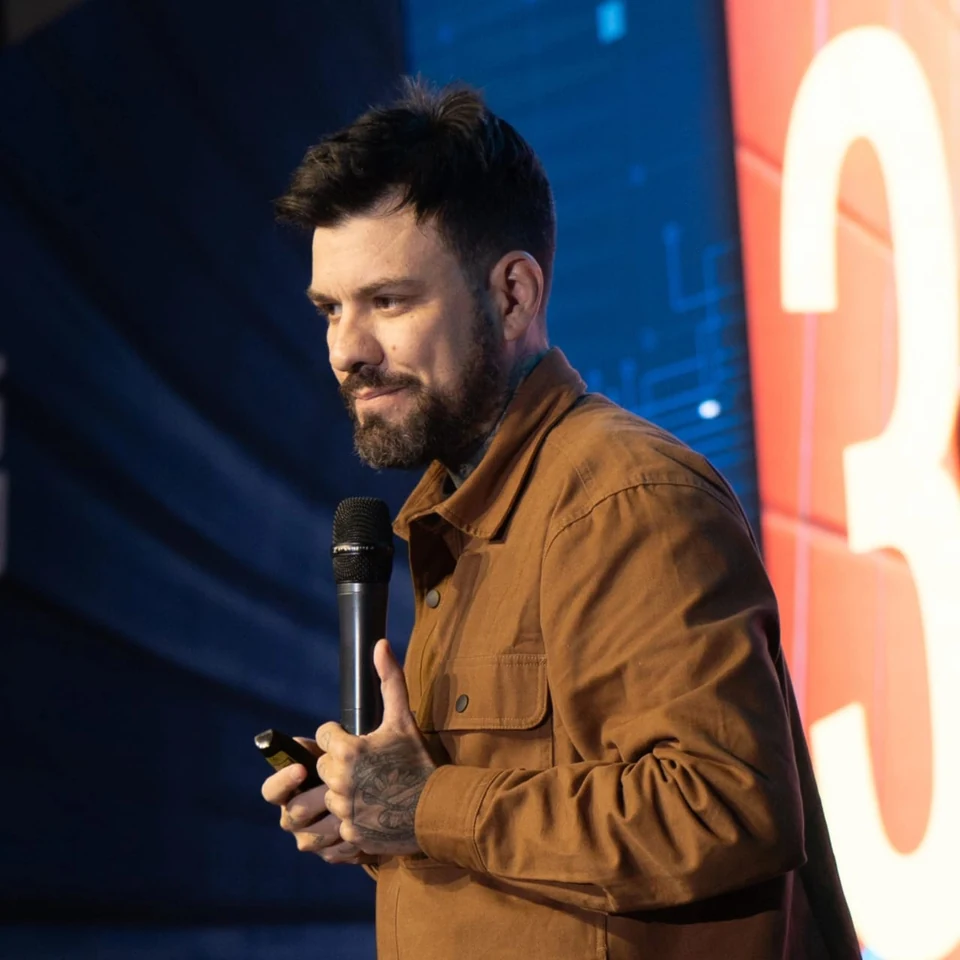 Gui Zanoni no palco, de jaqueta marrom, com o microfone e o passador de slides na mão, diante de um telão azul.