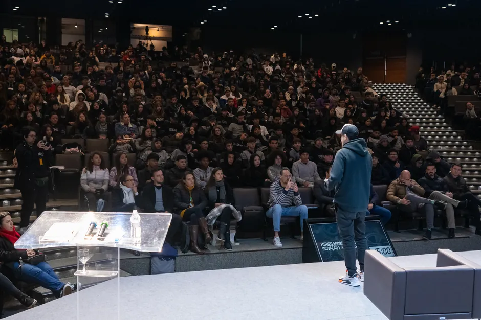 Auditório lotado visto do palco. No chão, o monitor de retorno mostra o relógio do palestrante marcando 15:00.