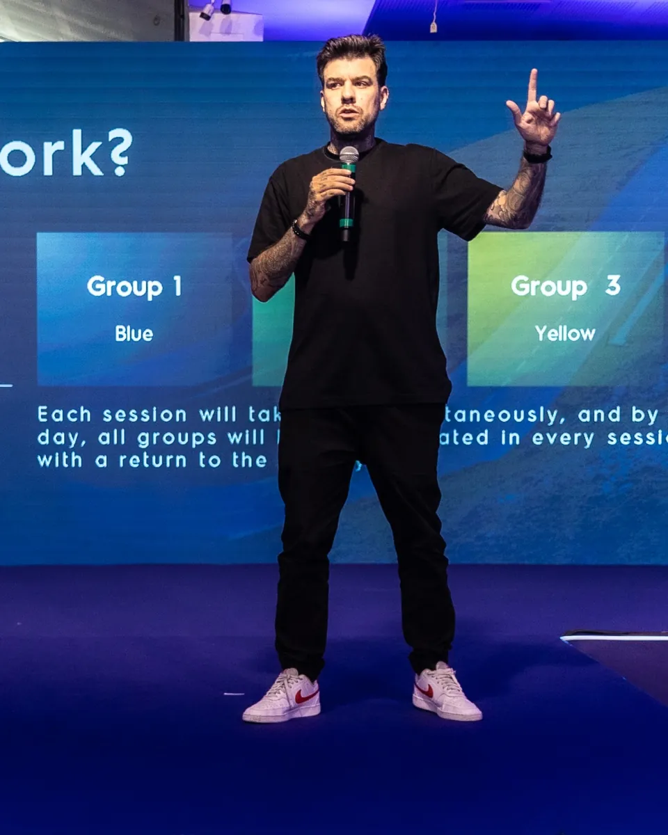 Gui Zanoni no palco, de camiseta preta e microfone na mão, com o dedo para o alto. No telão atrás dele, a divisão da sala em grupos por cor: Group 1 Blue, Group 3 Yellow.