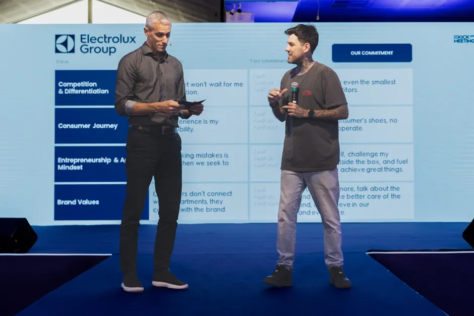 Gui Zanoni e um executivo no palco, diante de um telão da Electrolux Group com uma tabela: os valores da empresa numa coluna e, ao lado, frases de compromisso que começam com I will.