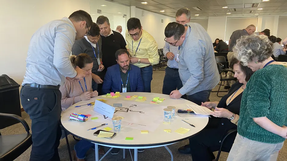Um grupo de profissionais em volta de uma mesa redonda coberta de post-its coloridos, canetas e uma folha grande com desenhos. Uma placa sobre a mesa indica o número dela.