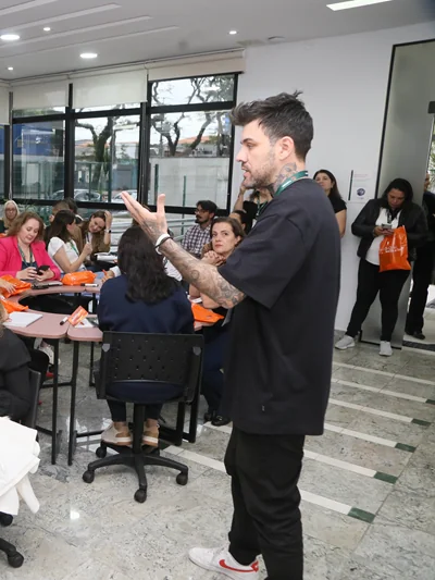 Gui Zanoni de pé, gesticulando, ao lado de participantes sentados em mesas durante um workshop.