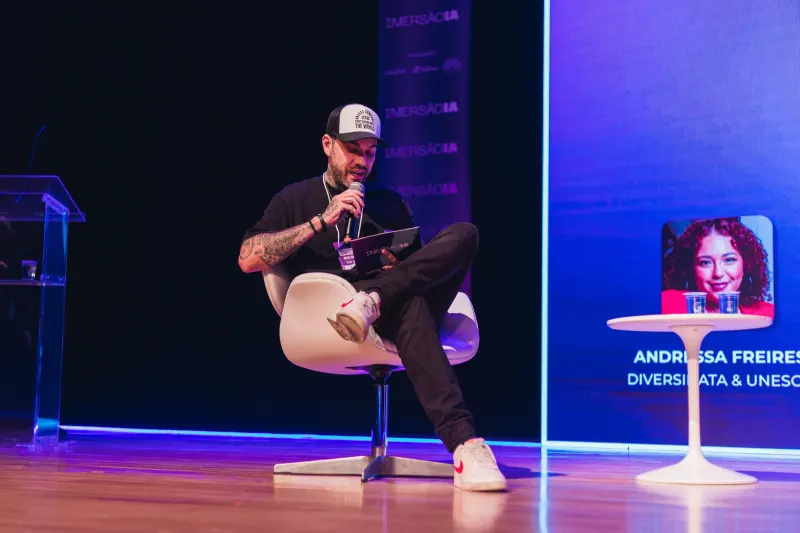 Gui Zanoni sentado numa poltrona branca no palco, numa conversa de evento