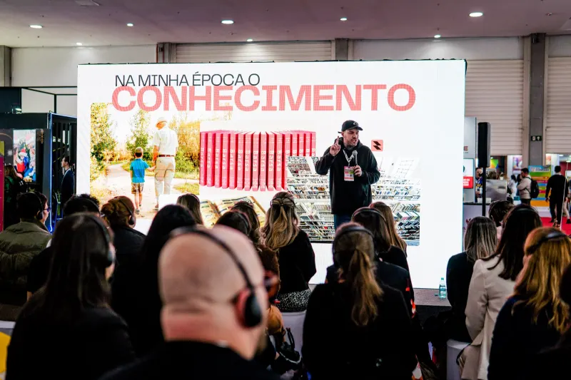 Gui Zanoni ao microfone num estande de congresso, diante da plateia, com o telão
