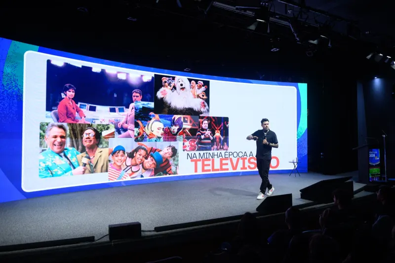 Gui Zanoni num palco grande com telão curvo, mostrando programas antigos de televisão